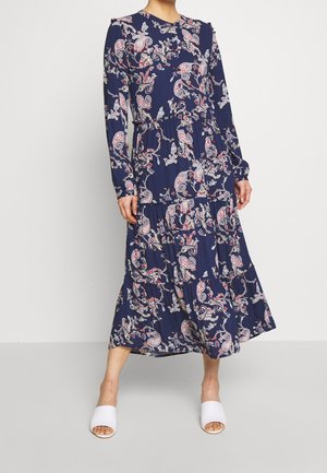 Kobieta ubrana w granatową, długorękawową sukienkę midi z różowo-białymi wzorami paisley oraz białe, otwarte buty na stopę.