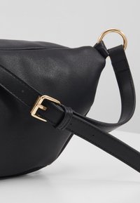 Musta nahkainen käsilaukku pyöreällä muotoilulla, jossa on kultainen metallirengas ja solki säädettävässä hihnassa. Suora pinta.