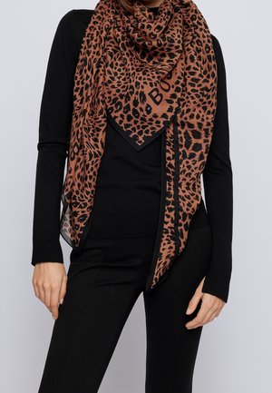 Femme portant un haut noir à manches longues et un pantalon, avec une grande écharpe marron et noire à imprimé léopard drapée autour du cou.