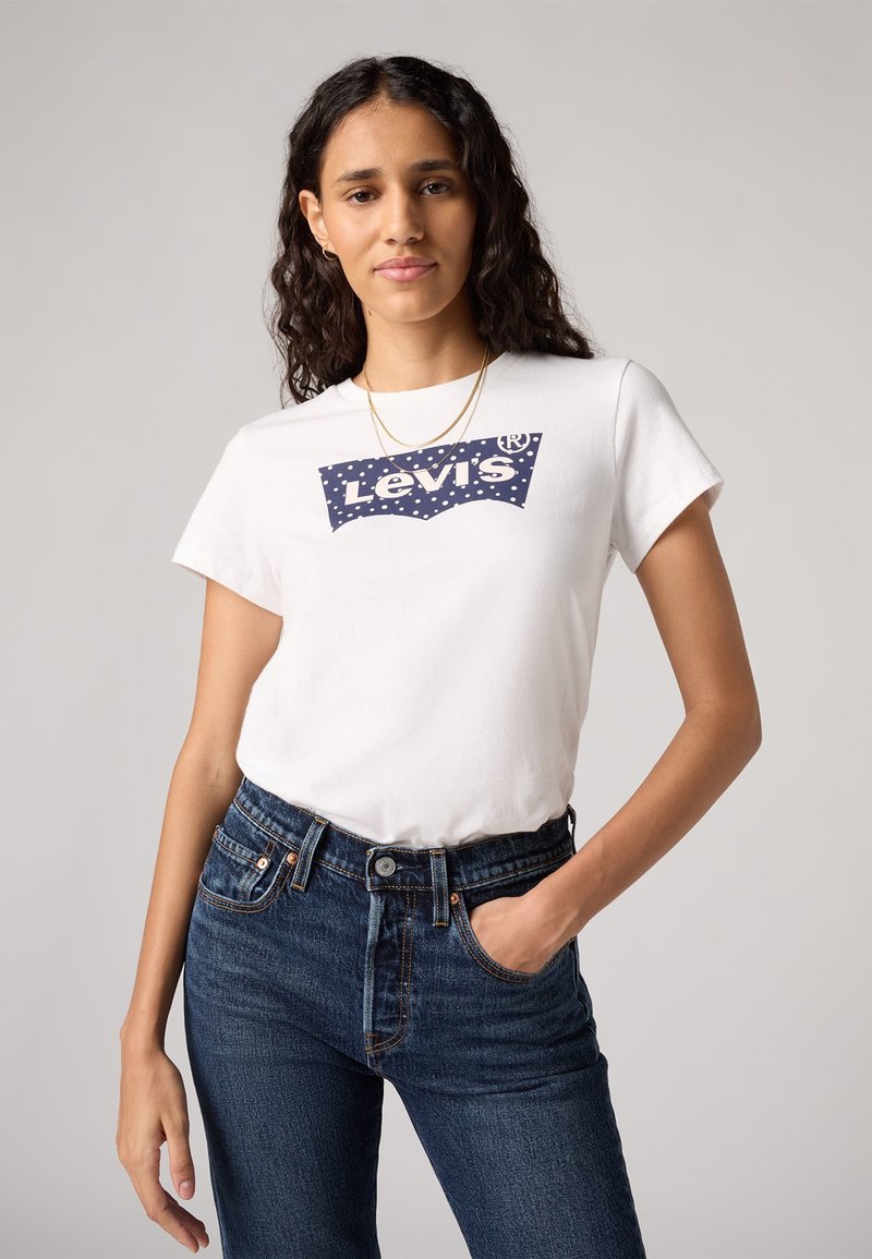 Camiseta blanca de algodón con un logo de Levi's azul y un diseño de costura curva, combinada con unos jeans de mezclilla azul oscuro.