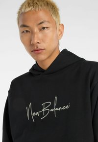 Sudadera negra de tela con un gran logo en cursiva blanca de "New Balance" en el frente. El diseño incluye una capucha con cordón.