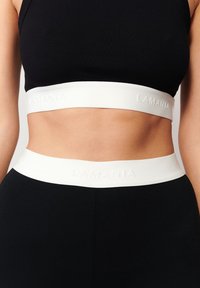 Czarny top crop z białą, wypukłą taśmą w pasie z napisem "LAMANIA". Gładki materiał, dopasowany krój, podkreślający środkową część ciała.
