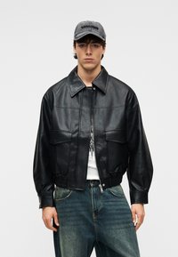 Μαύρο δερμάτινο μπουφάν τύπου bomber με γιακά, κλείσιμο μπροστά με φερμουάρ και δύο τσέπες στο στήθος. Φορεμένο πάνω από λευκό πουλόβερ με γράμματα κα ι μπλε τζιν.