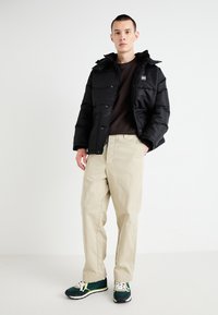 Black puffer jacket with a hood, beige trousers, and green sneakers. The jacket features a prominent logo and snap-button closure.