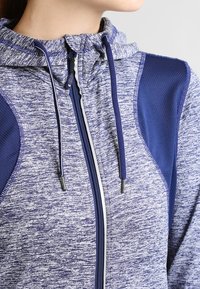 Sudadera con cremallera que presenta un patrón texturizado en azul y gris, con paneles en azul contrastantes, cordones y un cuello alto para mayor protección.