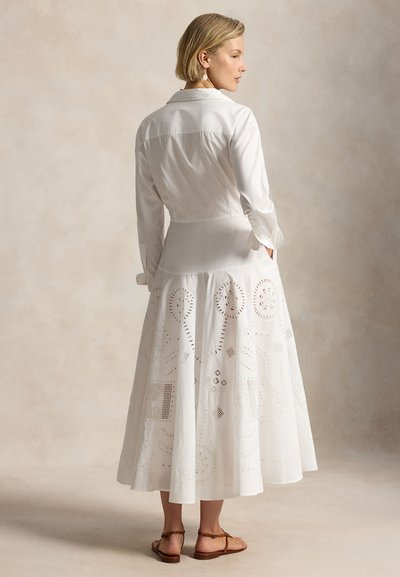 Femeie cu păr scurt blond, purtând o rochie albă lungă cu modele decupate și sandale maro plate, stând pe un fundal simplu.