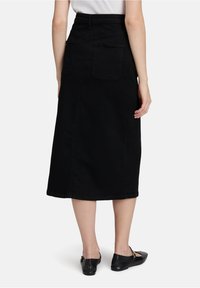 Femme portant une jupe midi noire taille haute avec des poches arrière et des ballerines noires type Mary Jane, se tenant devant un fond blanc.