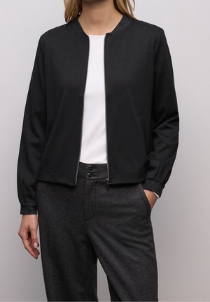 Femme portant une veste noire zippée avec col et poignets côtelés, chemise blanche, pantalon gris foncé taille haute avec boutons, main dans la poche.