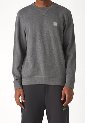 Homme portant un sweat-shirt gris uni à manches longues et col rond avec un petit logo BOSS sur la poitrine, ainsi qu'un pantalon foncé avec un logo BOSS.