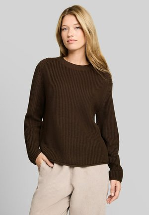 Junge Frau mit langen blonden Haaren, die einen dunkelbraunen gestrickten Pullover und hellbeige Hosen trägt, steht mit einer Hand in der Tasche.
