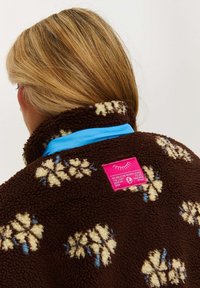 Hnědá fleecová bunda s krémovými a modrými květinovými vzory, vysokým límcem a růžovou etiketou umístěnou na zadní straně. Viditelná modrá vnitřní podšívka.