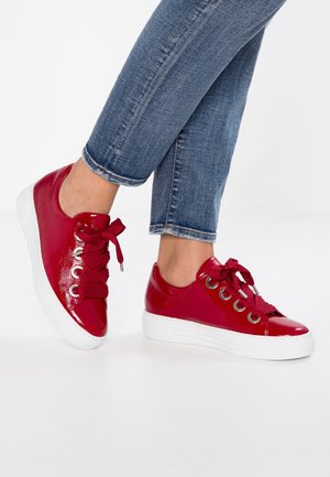 Persona con vaqueros azules y zapatillas plataforma rojas brillantes con cordones gruesos y ojales metálicos, de pie sobre suelo blanco.