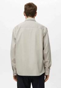 Mężczyzna z kr�ótkimi brązowymi włosami, w jasnobeżowej koszuli z długim rękawem i ciemnych spodniach, widziany z tyłu na jednolitym białym tle.