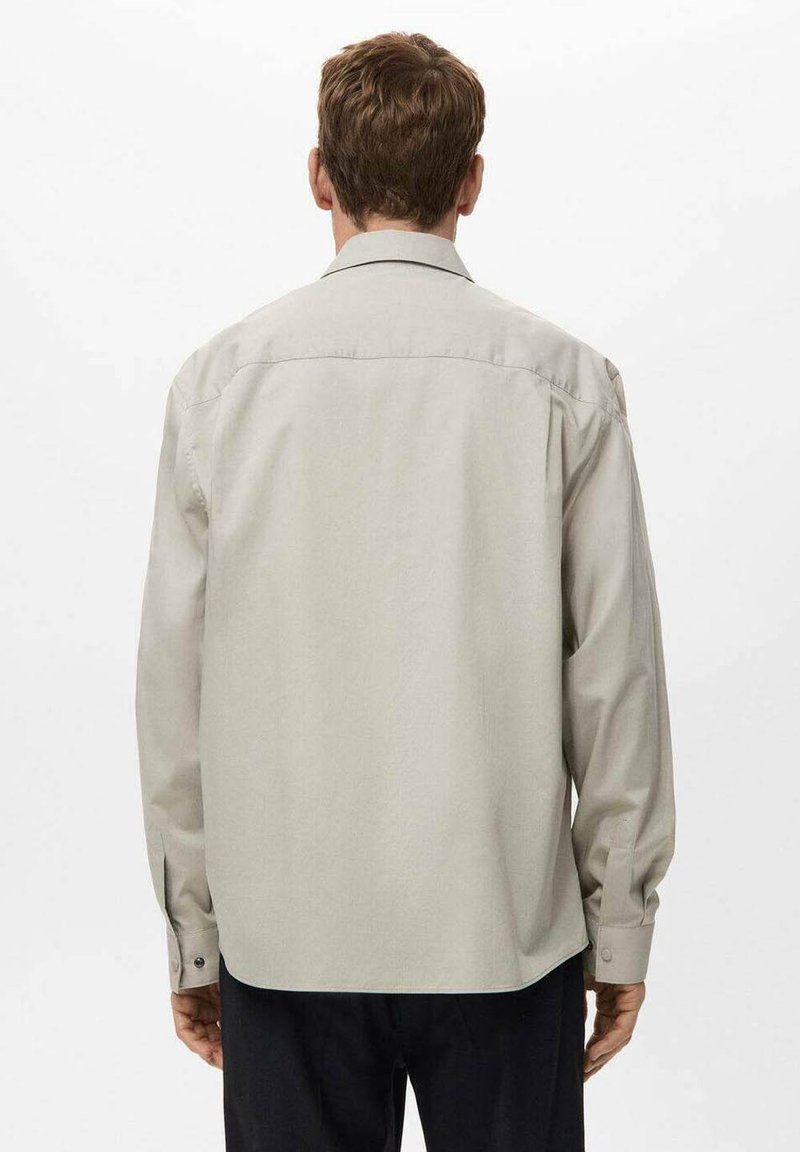 Ein Mann mit kurzen braunen Haaren, der ein hellbeiges Langarmhemd und dunkle Hosen trägt, von hinten vor einem schlichten weißen Hintergrund betrachtet.