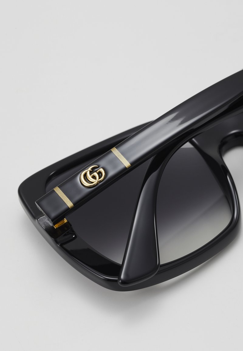 Black sunglasses with a wide, angular frame, featuring gradient dark lenses. Gold accents on the temples display a double "G" logo.