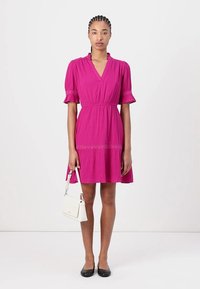 Fuchsia jurk met korte mouwen en een V-hals, met een rimpelrand en gedetailleerd met horizontale stiksels. Model houdt een kleine witte tas vast.