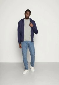 Joven vestido con sudadera navy, camiseta gris, vaqueros azules y zapatillas blancas, de pie contra un fondo claro y liso.