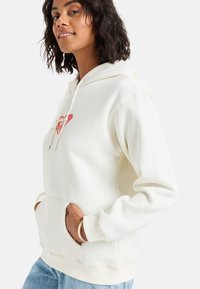 Sudadera color crema con un bolsillo frontal, capucha con cordones y un diseño sutil de logo en rosa y dorado. Tejido suave con puños acanalados.