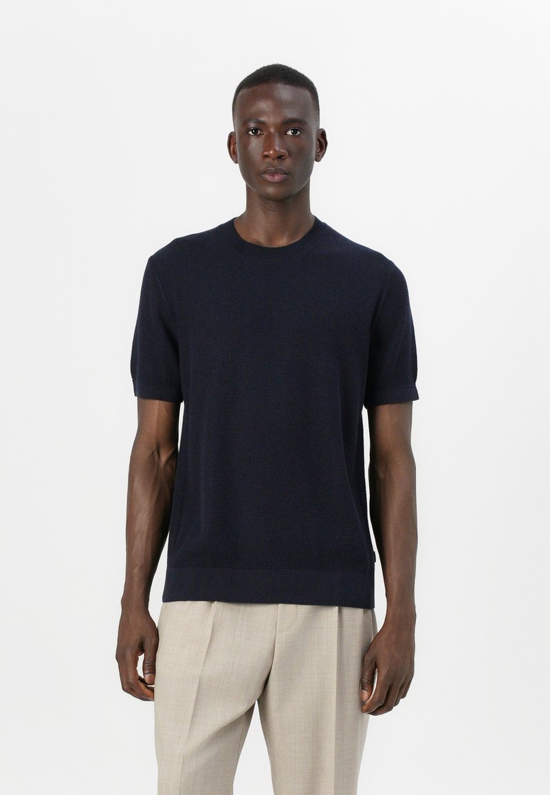 Mornarsko modra pletena majica s kratkimi rokavi, okroglo ovratnico in teksturirano tkanino. Kombinirana s svetlo bež, nabranimi hlačami.