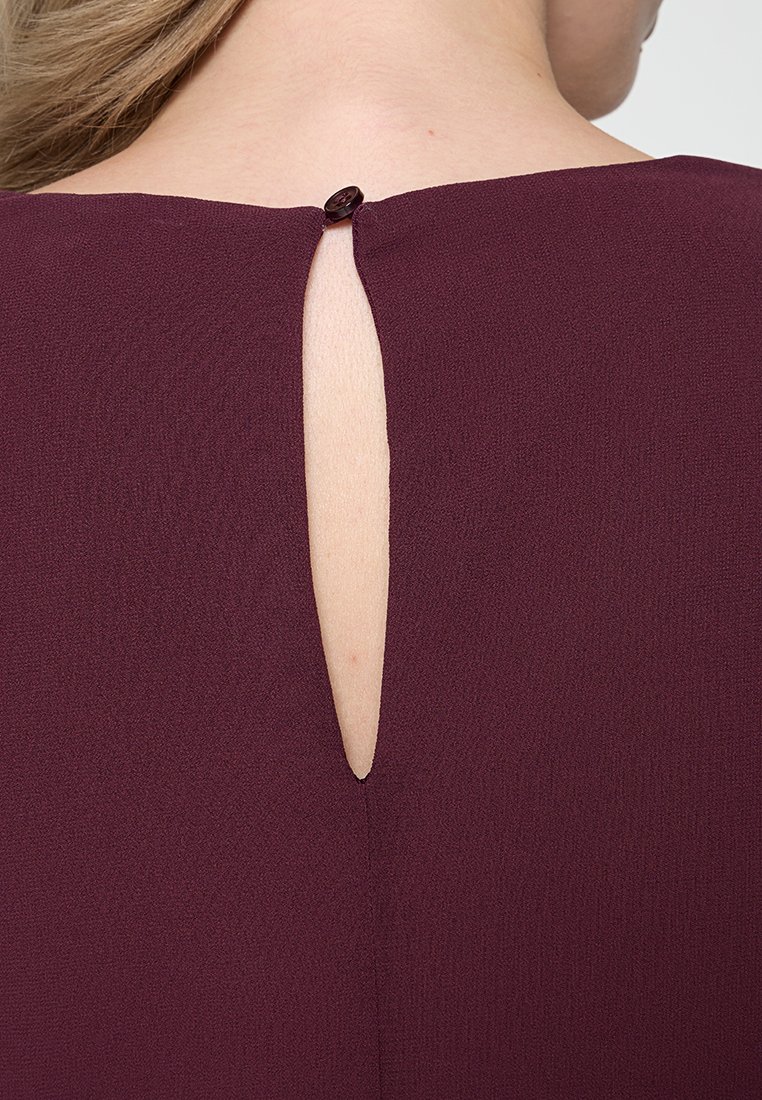 Bordeauxrotes Kleid mit einer Schlüsselöffnung hinten, die mit einem runden Knopf gesichert ist. Weicher, strukturierter Stoff, der ein einfaches und klares Design aufweist.