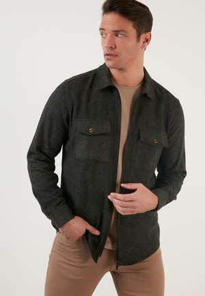 Dark green, textured fabric shirt jacket with two chest pockets and a zip front. Features a classic collar and fitted sleeves.