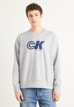 Grå hettegenser laget av bomullsblanding, med en stor marineblå logo "CK" og teksten "Calvin Klein". Myk tekstur, ribbede ermer og nederkant.