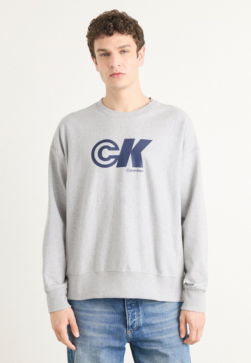 Šedá mikina vyrobená z bavlněné směsi, s velkým námořnickým logem "CK" a textem "Calvin Klein". Měkká textura, žebrované manžety a lem.