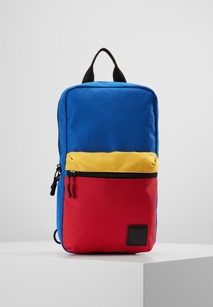 Sac à dos rectangulaire avec corps principal bleu, poche centrale zippée jaune et poche inférieure rouge, posé sur une surface blanche.