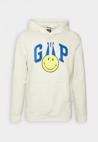 Sudadera crema hecha de algodón, con un bolsillo delantero, cordones blancos, letras GAP en azul y un gran gráfico de una cara sonriente amarilla.