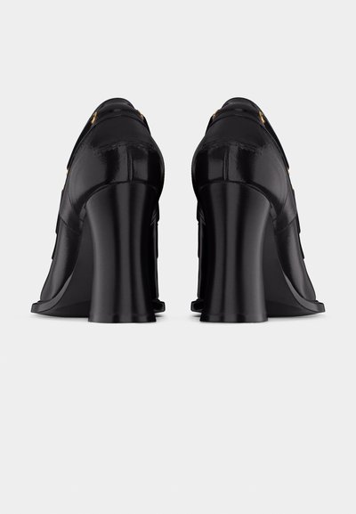 Ζευγάρι μαύρα δερμάτινα παπούτσια με ψηλό τακούνι και χοντρό τετράγωνο τακούνι με χρυσές αγκράφες, εμφανιζόμενα από πίσω σε απλό φωτεινό φόντο.