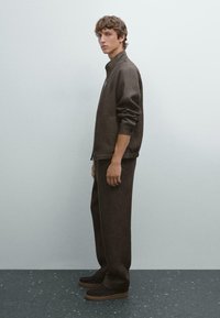 Brown linen jacket with high collar and long sleeves, paired with matching brown trousers and dark sneakers with a thick sole.