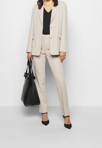 Blazer beige con botones dorados, combinado con pantalones beige a medida. Top y bolso negros. Tacones negros de punta. Diseño suave y limpio.