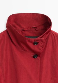 Veste rouge avec un col montant fermé par deux boutons noirs, présentant une texture lisse et une boucle de suspension cachée à l'intérieur du col.