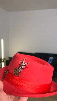 Rood vilten fedora-hoed met een brede lintband. Heeft een decoratieve veer en metalen accent aan de zijkant. Gladde textuur, gestructureerde vorm.