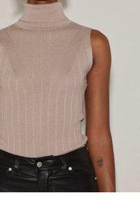 Beige ribbad polokrage utan ärmar med vertikala linjer och en V-formad detalj, kombinerad med höga svarta byxor och en silverfärgad knapp.