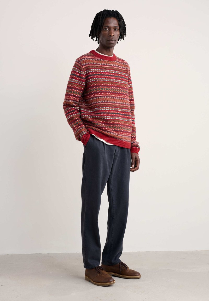 Sarkans džemperis ar ģeometriskām apdrukām, ar ribotām piedurknēm un kaklu. Valkāts ar tumši pelēkiem biksēm un brūnām šņorējamām kurpēm.