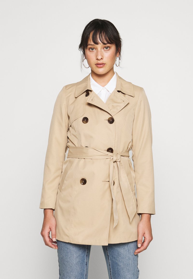 Beige trench coat s dvojitým zapínáním, páskem v pase, klopovým límcem a hladkým materiálem. Nosit s bílou košilí a džíny.