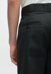 Sorte bukser med en glat tekstur, der har en baglomme med knap og en flad talje. Synlig syning på stoffet.