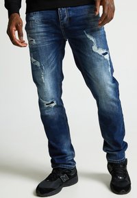 Jeans de mezclilla en azul oscuro con áreas desgastadas y desgarrones visibles. Diseño ajustado con una pierna ligeramente acampanada, combinados con zapatillas deportivas negras.