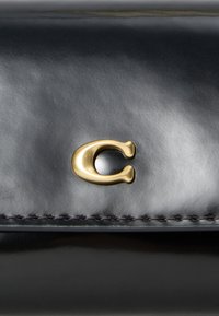 Crna kožna torba s zlatnim metalnim logom "C", glatke teksture i tamnim detaljima šavova uz rubove. Kompaktni i elegantni dizajn.