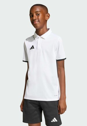 Chłopiec w białej koszulce polo Adidas i czarnych szortach Adidas, stojący z delikatnym uśmiechem na jednolitym jasnym tle.