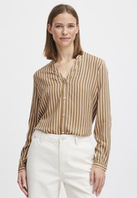 Brown striped blouse with a soft texture, featuring a V-neckline and button closure. Long sleeves have contrasting white cuffs. Worn with white pants.