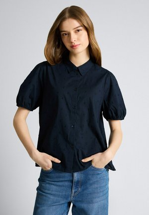 Młoda kobieta o jasnobrązowych włosach, ubrana w czarną koszulę na krótki rękaw z fakturą i niebieskie dżinsy, ręce w kieszeniach, neutralne tło.