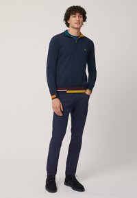 Pullover blu navy con colletto a zip, caratterizzato da accenti a righe multicolori sull'orlo e sui polsini. Indossato con pantaloni blu fitted e scarpe nere.