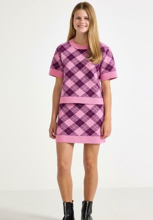 Jonge vrouw glimlacht, draagt een roze en paarse argyle korte mouw trui en bijpassende rok met zwarte enkellaarsjes op een witte achtergrond.