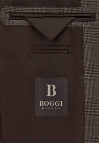 Tessuto marrone con una texture a motivi; presenta un'etichetta grigia con un logo e il testo "BOGGI MILANO"; include una tasca e dettagli.