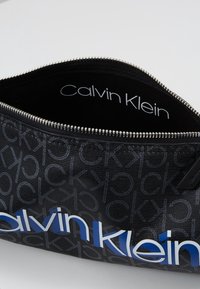 Borsa cosmetica nera con motivo ripetuto "CK". Presenta una cerniera argento e un logo "Calvin Klein" blu sulla parte interna anteriore. Tessuto strutturato.
