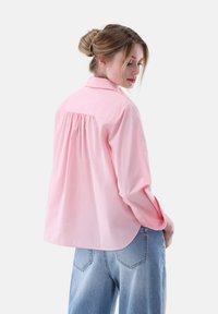 Light pink button-up shirt with a relaxed fit, featuring a back pleat and long sleeves, paired with high-waisted blue jeans.