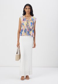 Farbige, durchscheinende Bluse mit einem Peplum-Saum und Schnürung vorne. Kombiniert mit einem strukturierten weißen Maxikleid und neutralen Sandalen. Hält eine geflochtene Tasche.