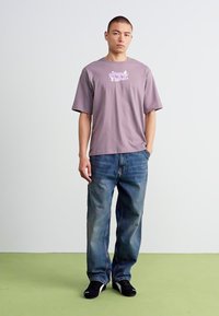 Mand står med ryggen mod en hvid væg iført en lilla grafisk T-shirt, blå jeans og sorte sko med den ene hånd i lommen.